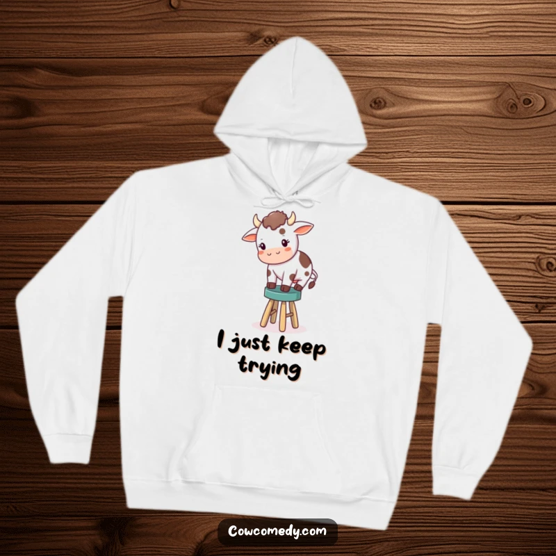 Funny hoodie design of a small cow wobbling precariously while trying to maintain balance on an extremely tiny stool, conveying adorable awkwardness.